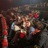 Grupa uczniów z klasy siedzi w kinie, oczekując na rozpoczęcie seansu filmowego.
